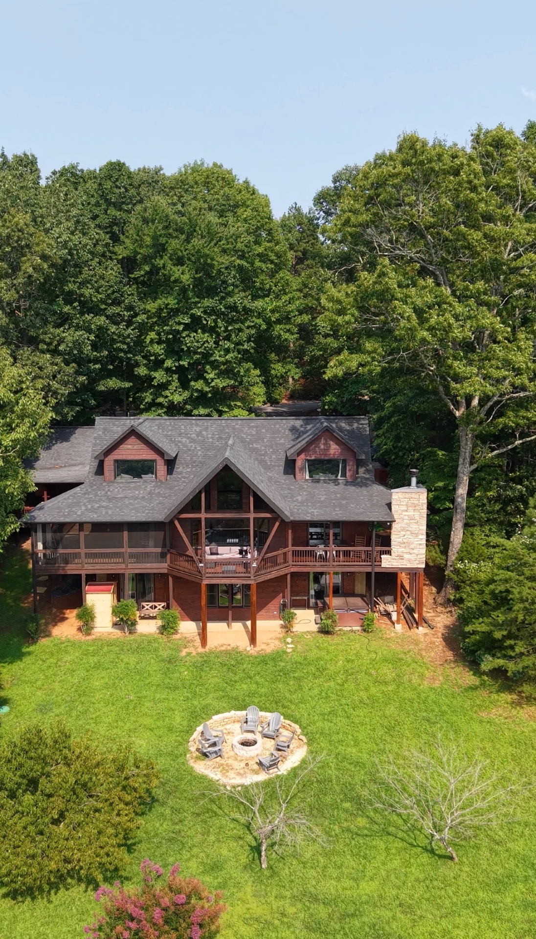 Aerial drone photograph of a timber lodge surrounded by dense forest, with a mown lawn and stone fire pit below it