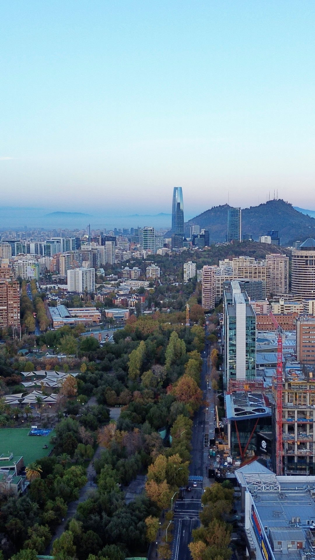 Aerial drone photograph of a city skyline at dawn with a green park and tree-lined avenue leading toward the towers