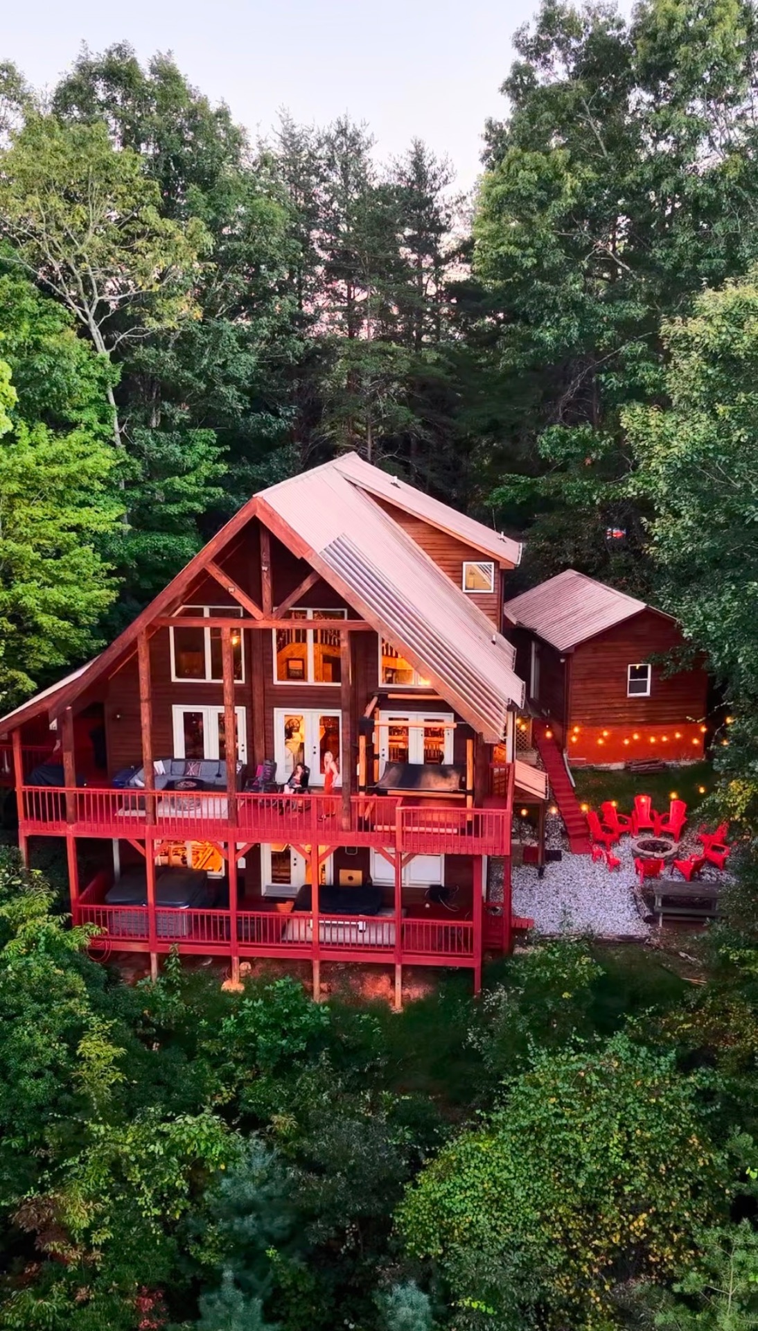 Aerial drone photograph of a timber lodge with red wraparound decks glowing at dusk, surrounded by forest, with a lit fire pit seating area beside it